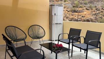 A covered outdoor seating area with two chairs and a table, a refrigerator in the background, and a view of a rocky landscape.
