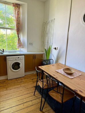 A compact kitchen with a washing machine, wooden table, chairs, and a window with curtains. - Leafy Marchmont 2-Bed · Meadows · City Centre (Edinburgh)
