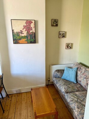 A room with a wooden floor, a sofa with patterned upholstery, a blue cushion, a painting of a landscape, and three small framed pictures on the wall. - Leafy Marchmont 2-Bed · Meadows · City Centre (Edinburgh)