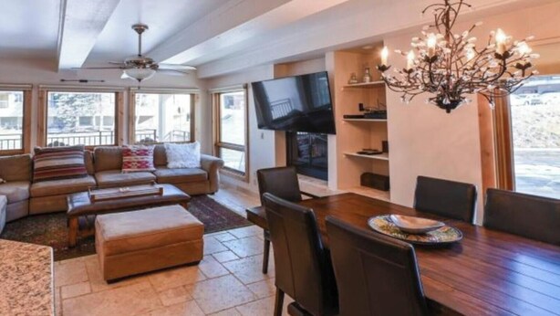 A spacious living room with a large sofa set, a dining table with chairs, a chandelier, and a TV mounted on the wall. - Ski Lodge Styled Plaza Condo 3 Bedroom Condo (Crested Butte)