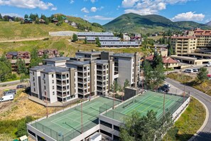 Un immeuble à plusieurs étages, doté d’un court de tennis sur le toit, entouré d’immeubles résidentiels et surplombant un paysage vallonné.