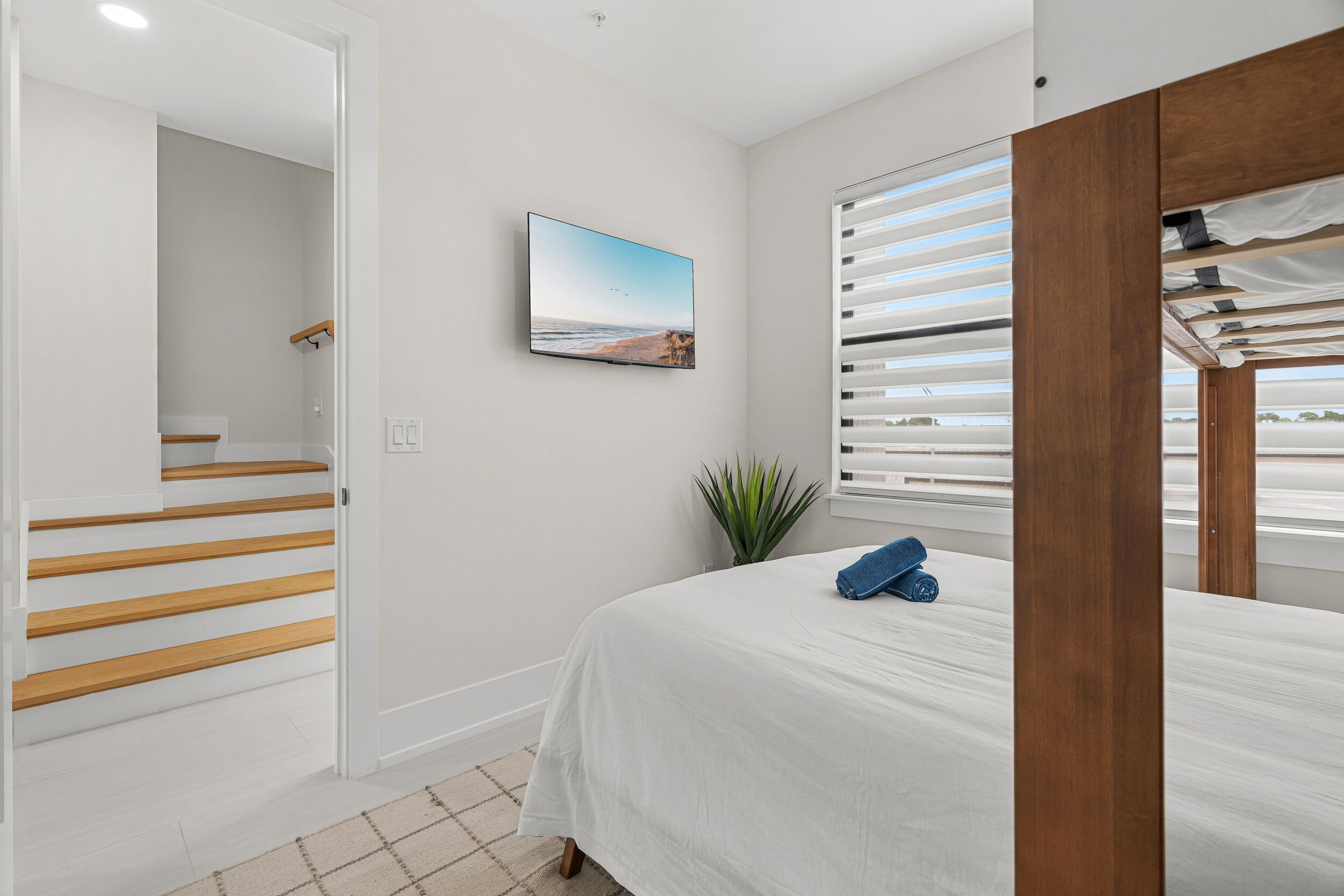 Een slaapkamer met een bed, een raam met zonwering, een plant en een aan de muur bevestigde televisie.