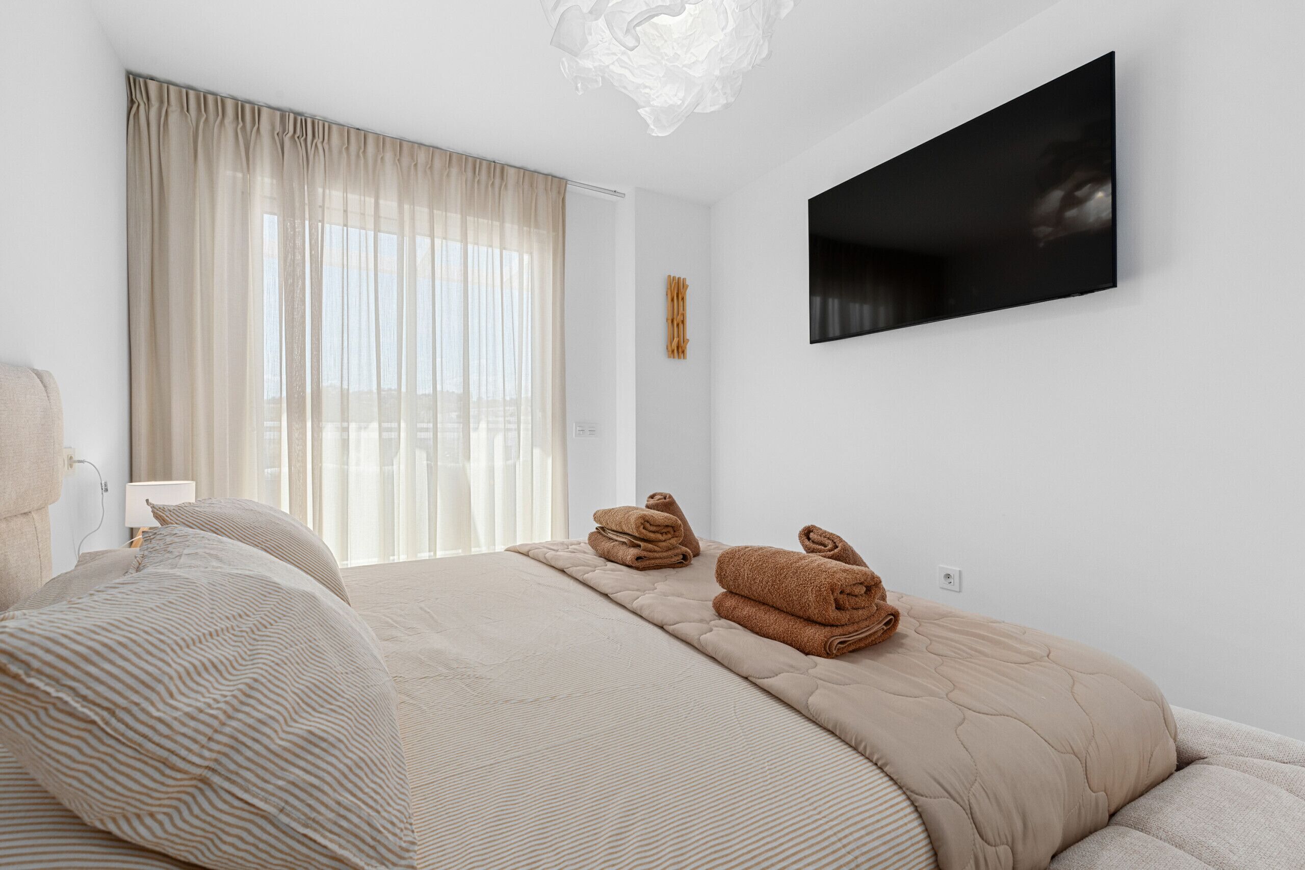 A hotel room with a bed, a flat-screen TV mounted on the wall, and a window with sheer curtains.