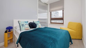 Un dormitorio con cama, mesita de noche, silla, ventana y lámpara colgante.