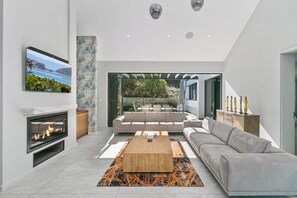 A modern living room with a fireplace, a large sofa, a coffee table, and a view of a garden through an open door. - Miles End @ Pezula (Knysna)