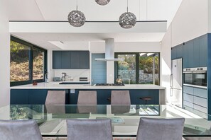 A modern kitchen with a central island, large windows, and a dining area with a glass table. - Miles End @ Pezula (Knysna)