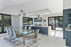 A modern kitchen with a central island, a dining area with a glass table and chairs, and a view of the outdoors. - Miles End @ Pezula (Knysna)