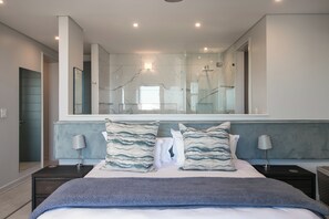 A modern bedroom with a large bed, two nightstands, a mirror, and a view of the bathroom. - Miles End @ Pezula (Knysna)