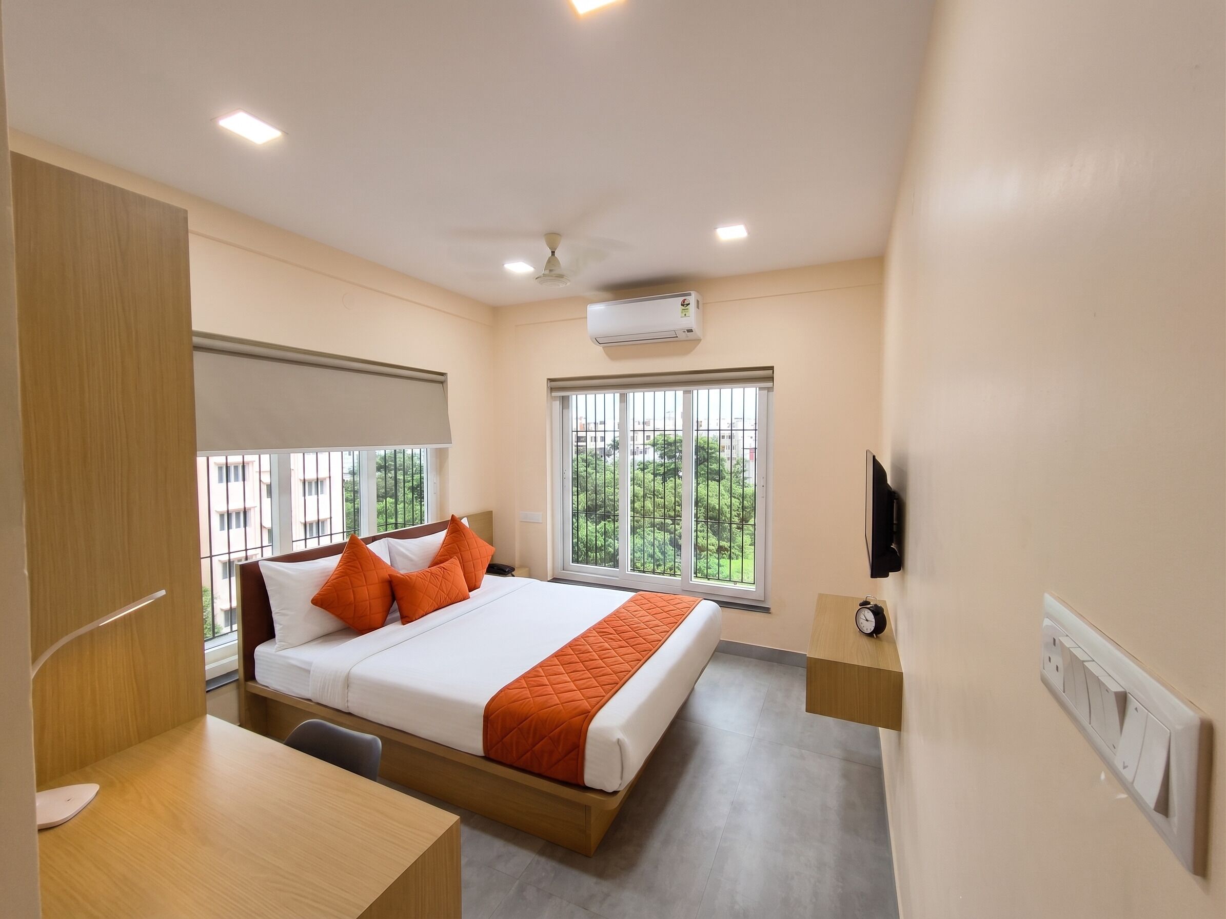 A hotel room with a bed, a desk, a chair, a TV, and a window with a view of greenery.