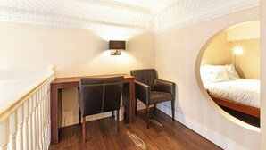 A hotel room with a wooden desk, two chairs, a bed with white linens, a wall-mounted lamp, and a circular window. - One Bedroom Duplex in Paddington (London)