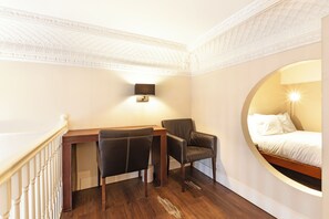 Ein Hotelzimmer mit Schreibtisch aus Holz, zwei Stühlen, einem Bett mit weißer Bettwäsche, einer Wandlampe und einem runden Fenster.