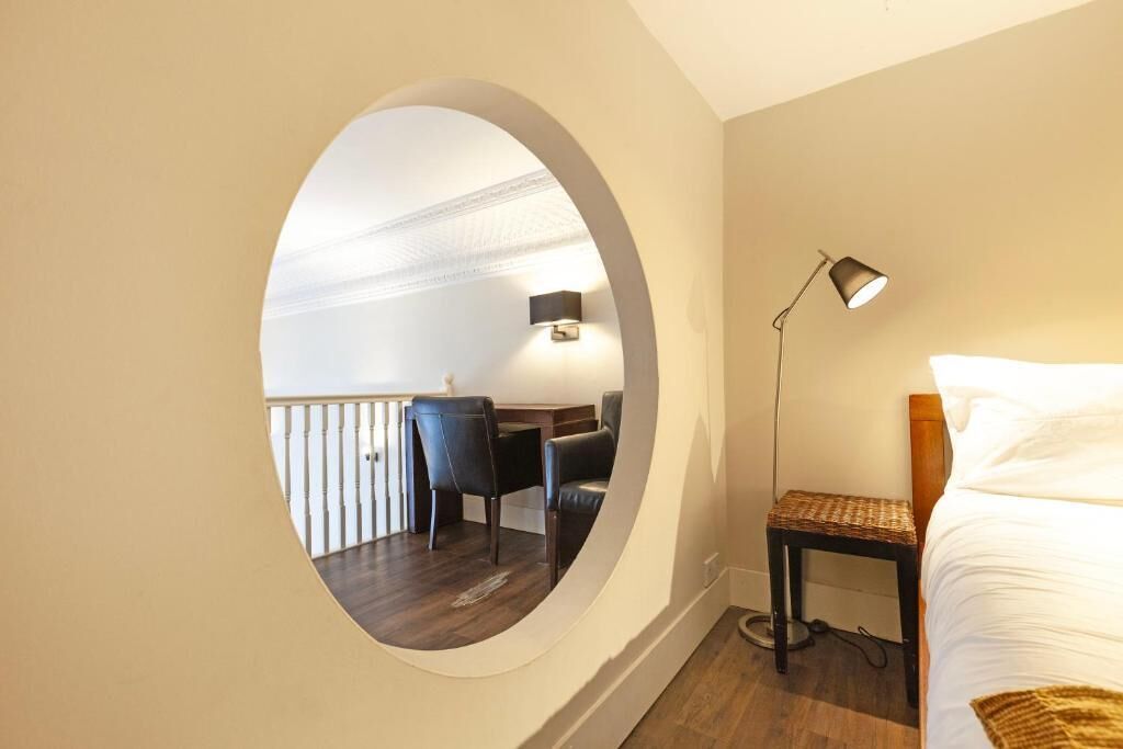 Quarto com cama, mesa de cabeceira, luminária e vista para uma varanda com mesa e cadeiras através de uma janela redonda.