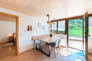 Ein Zimmer mit Holztisch, Stühlen, einer Bank, einer Pendelleuchte und einem Fenster mit Blick auf Grünflächen.