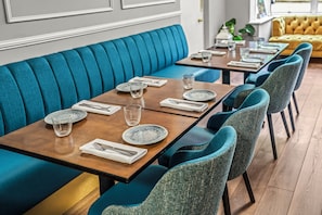 Un comedor con asientos tipo banca tapizados en color verde azulado y mesas de madera dispuestas con platos, vasos y cubiertos.