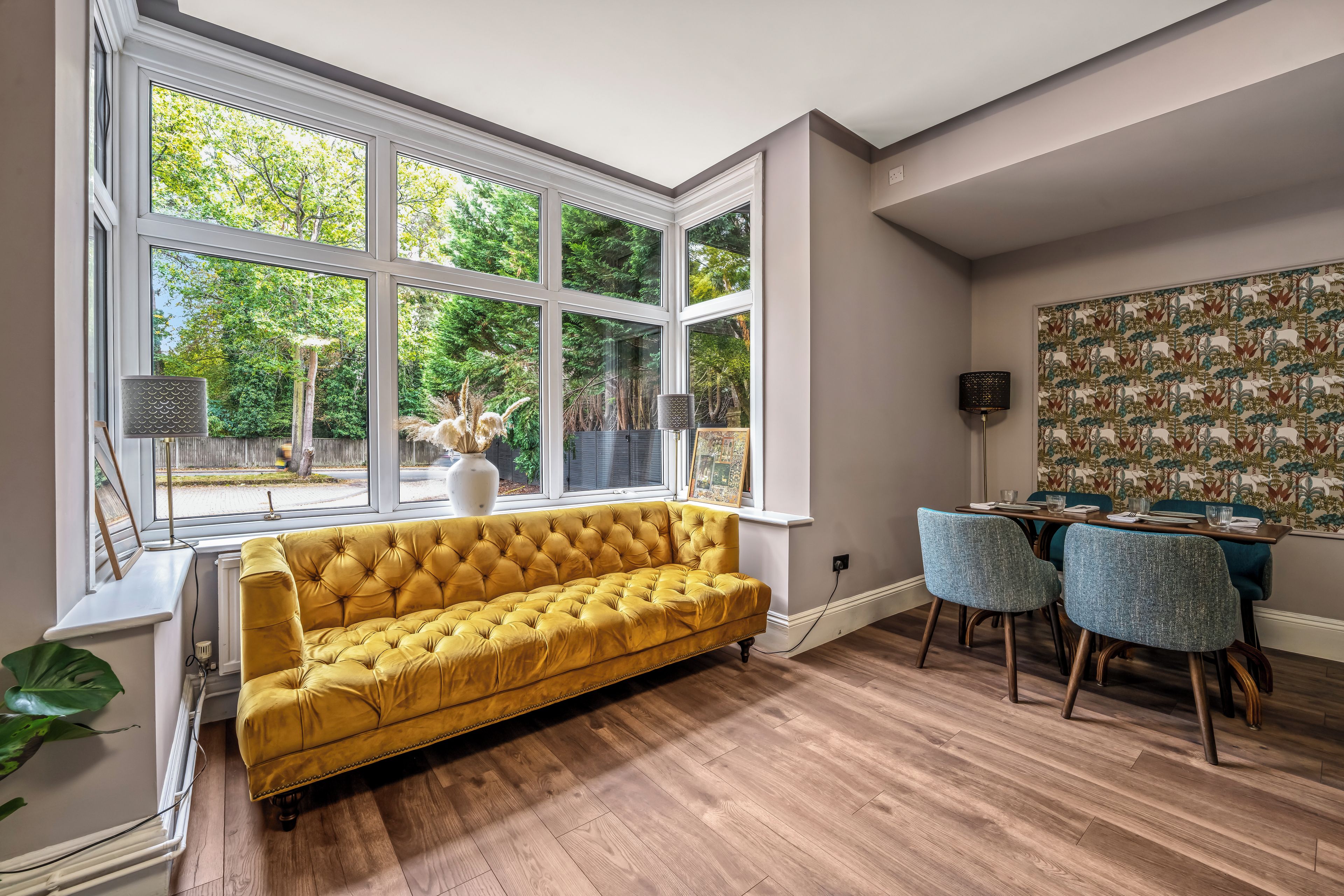 Une pièce avec un canapé jaune, une table à manger avec des chaises et de grandes fenêtres donnant sur la vue des arbres.