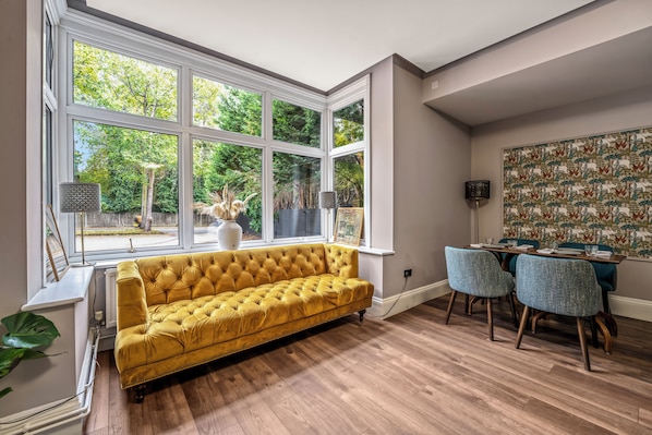 Una habitación con un sofá amarillo, una mesa de comedor con sillas y ventanas amplias con vista a árboles.