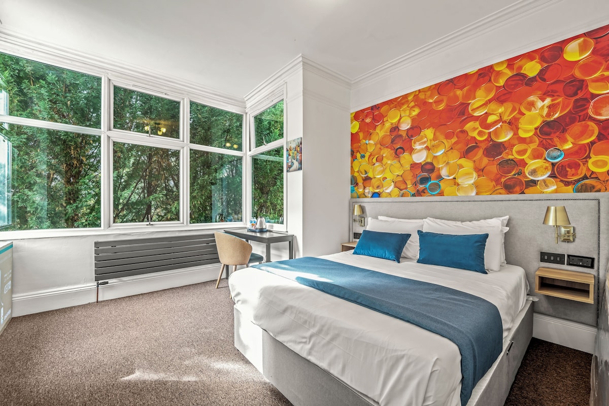 Una habitación de hotel moderna con una cama grande, un escritorio y vistas a zonas verdes a través de amplias ventanas.