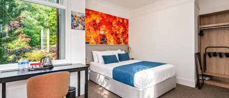 Habitación de hotel con cama, escritorio, silla, ventana con vista a follaje y una obra de arte colorida en la pared.
