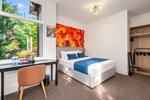 A hotel room with a bed, a desk, a chair, a window with a view of greenery, and a colorful artwork on the wall. - Seka Inn Hotel (Crowthorne)