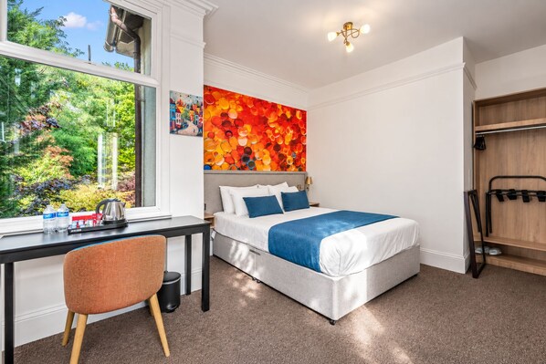 A hotel room with a bed, a desk, a chair, a window with a view of greenery, and a colorful artwork on the wall.
