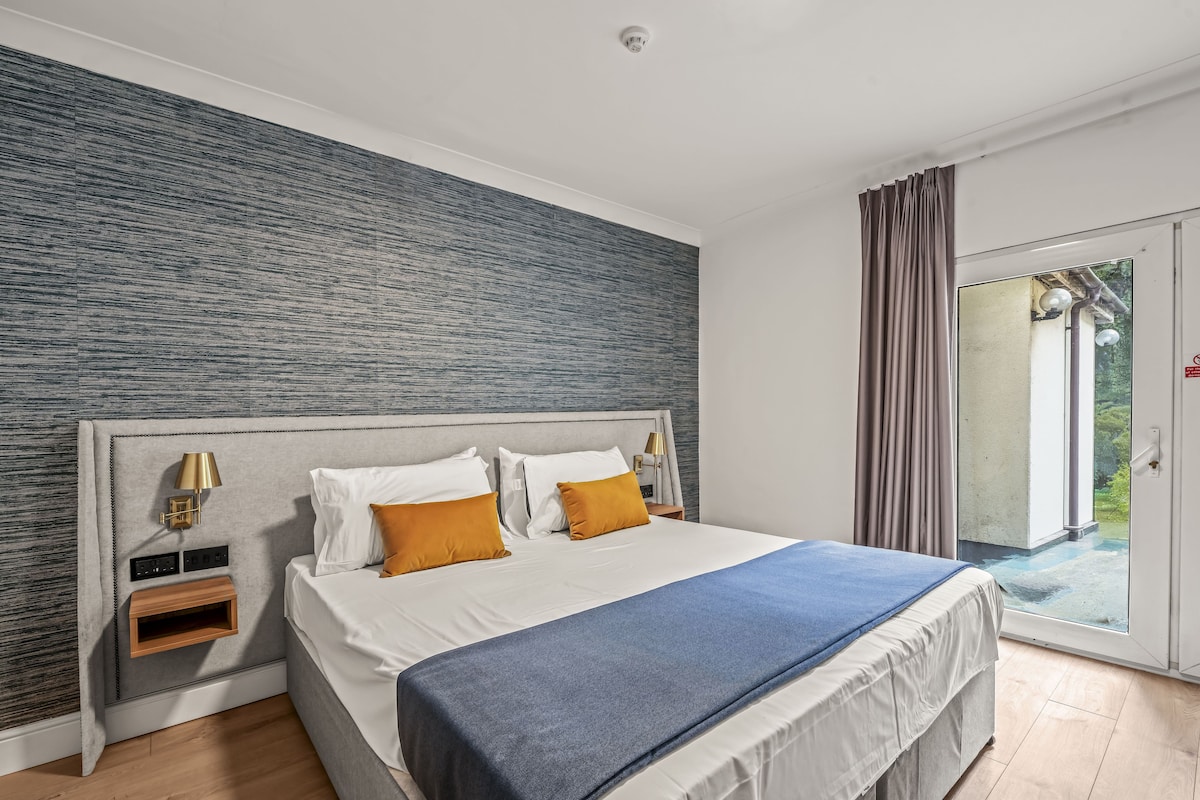 Habitación de hotel con cama, mesita de noche, lámpara y vistas al exterior.