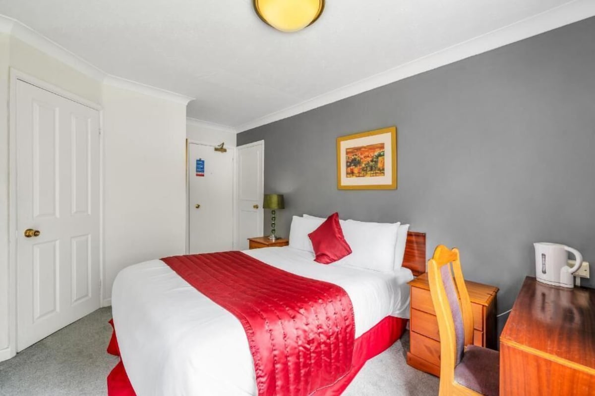 Una habitación de hotel con cama, escritorio, silla, mesita de noche y un cuadro en la pared.