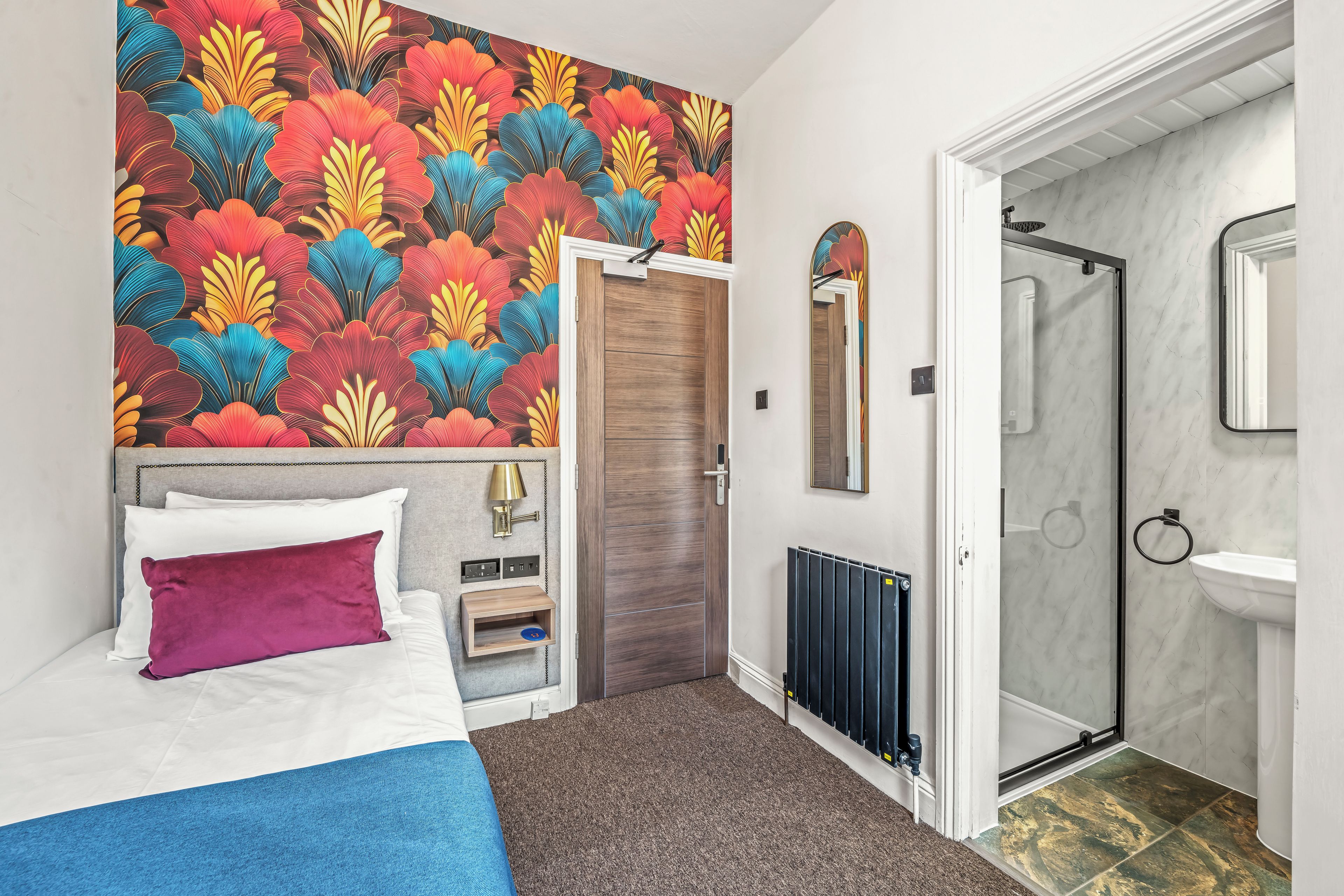 A hotel room with a bed, a shower, a toilet, and a wall with a colorful floral pattern.