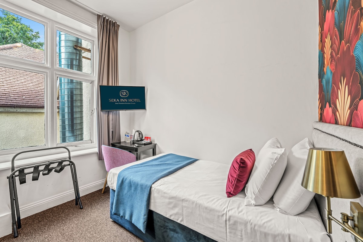 Habitación de hotel con cama, escritorio con tetera, silla, ventana con cortinas y un televisor de pared mostrando el nombre del hotel.