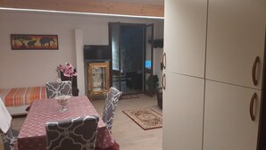 Ruokailutilassa on pöytä, tuoleja ja maljakko, jossa on kukkia. Taustalla näkyy sohva ja kaappi.