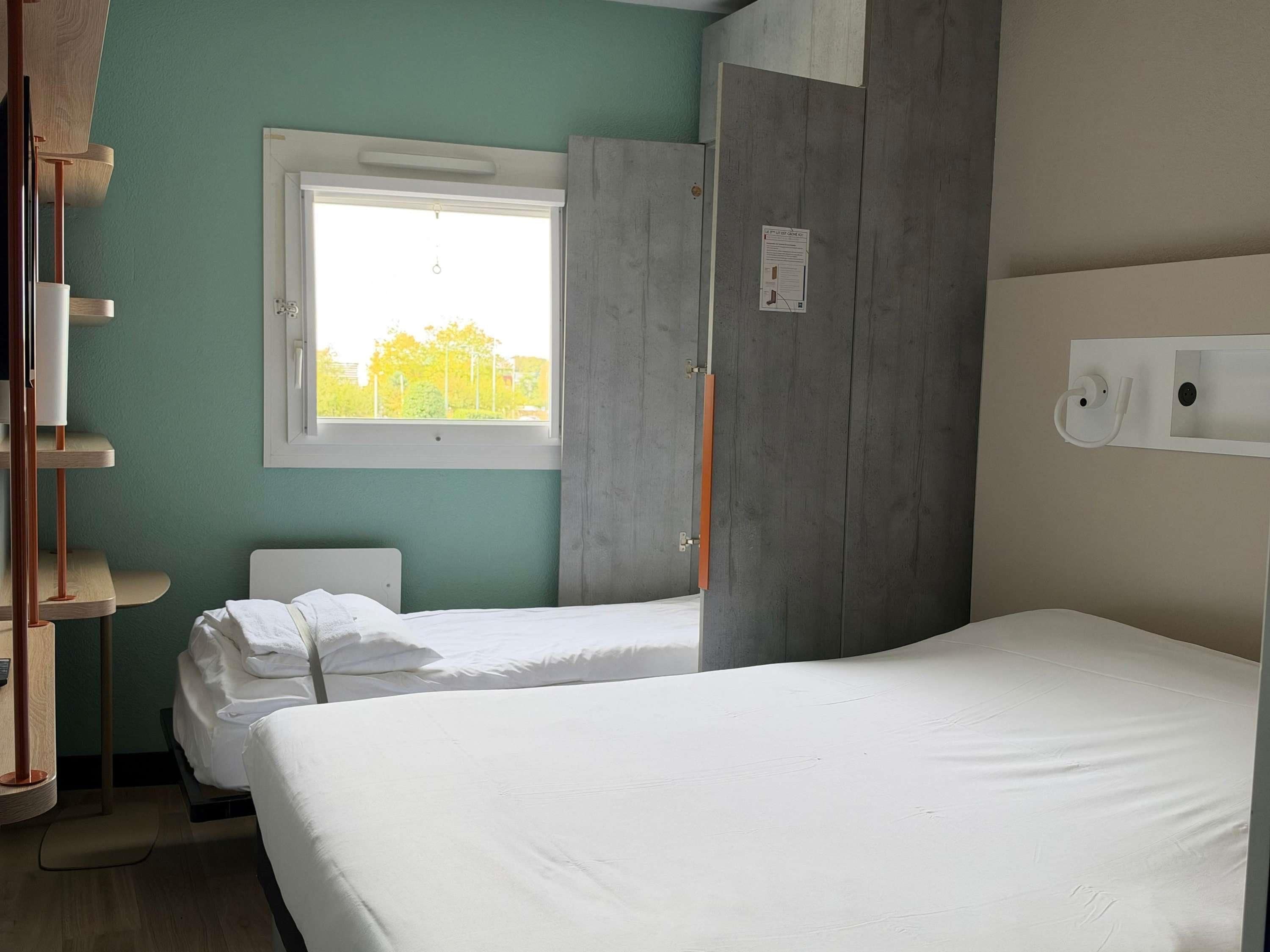 A hotel room with two beds, a window, a wooden wardrobe, and a wall-mounted light fixture.