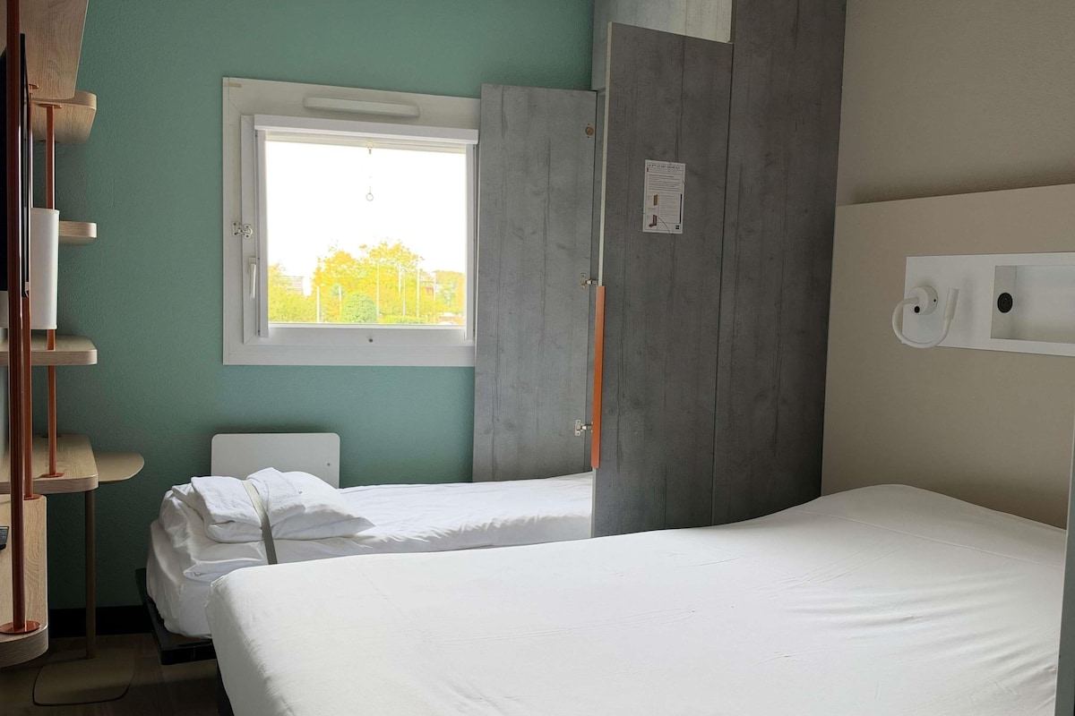 Habitación de hotel con dos camas, una ventana, un armario de madera y una lámpara fijada a la pared.