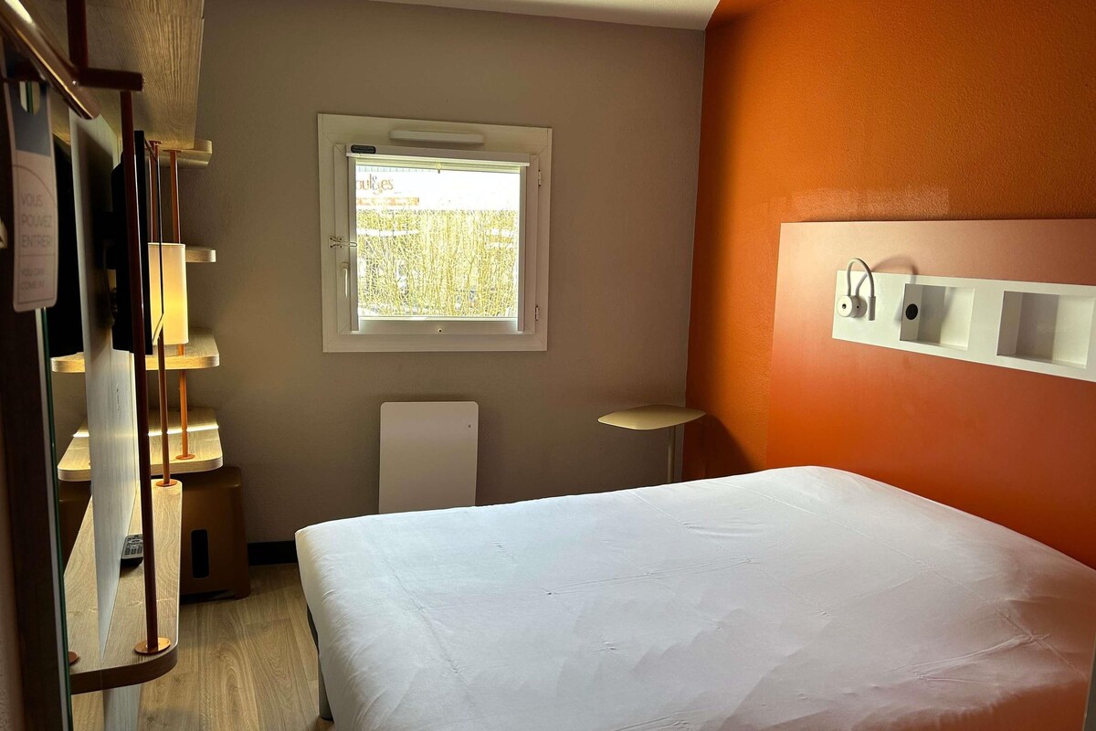 Een hotelkamer met een bed, een raam, een nachtkastje en een wandlamp.