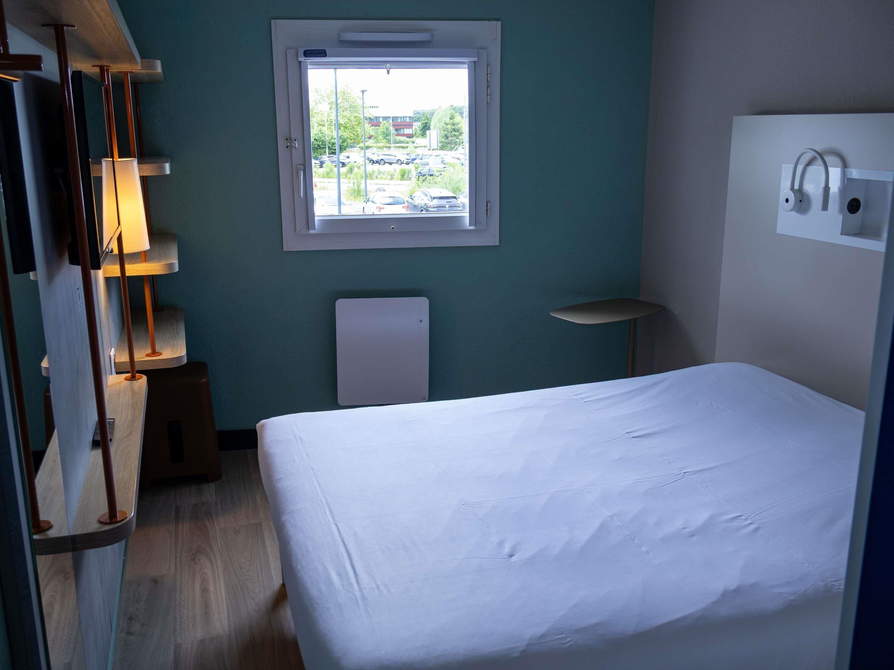 Una habitación de hotel con cama, ventana, mesita de noche y lámpara.