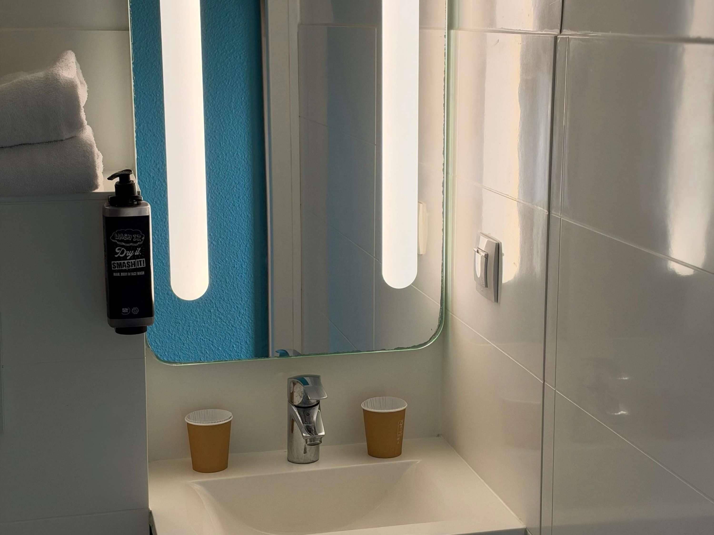 A bathroom with a mirror, a sink, two paper cups, a soap dispenser, and a towel.