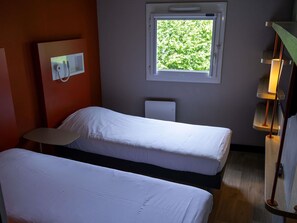 A room with two beds, a window with a view of greenery, a wall-mounted light, and a small table.