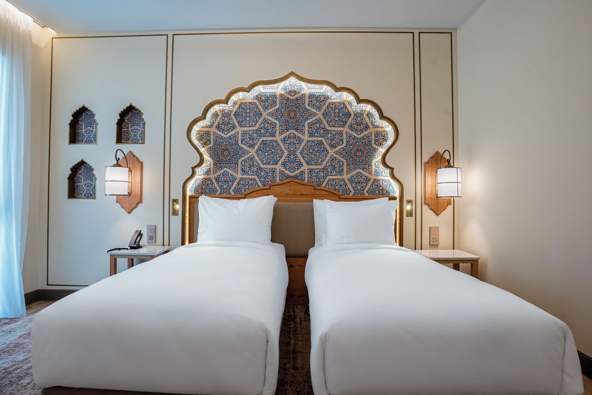 Una habitación de hotel con una cama grande, dos mesitas de noche, un cabecero decorativo y luces fijadas a la pared.