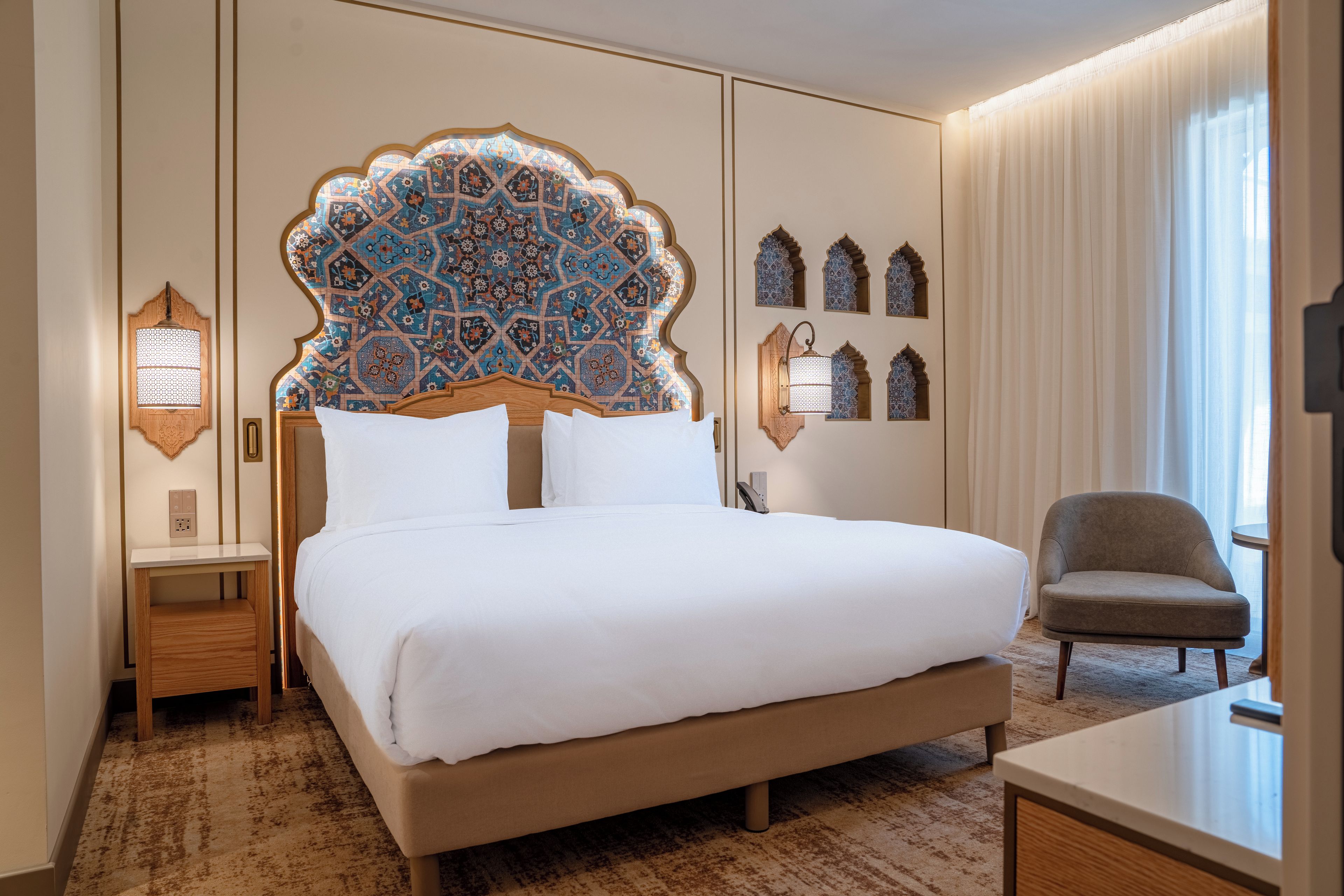 Quarto de hotel com uma cama grande, uma cadeira, uma escrivaninha e um painel decorativo com azulejos estampados.