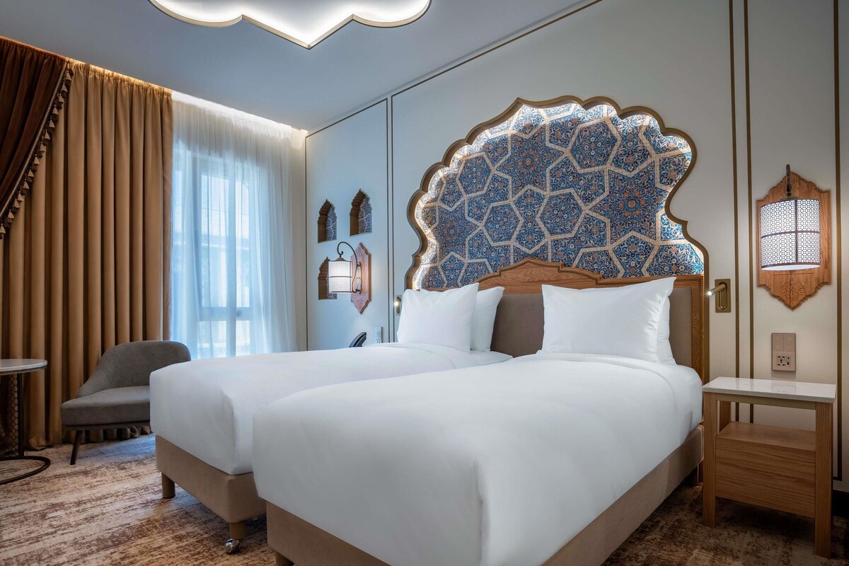 A hotel room with a large bed, a desk, a chair, and a window with curtains.
