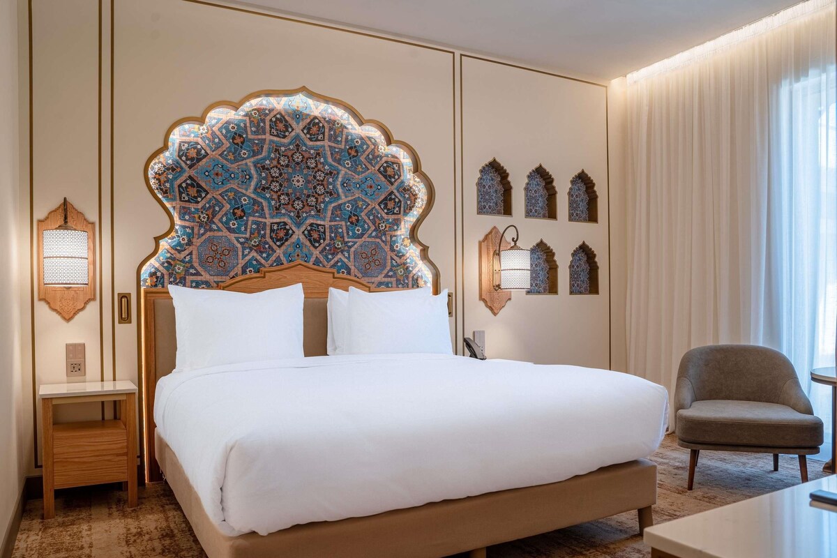 A hotel room with a large bed, a nightstand, a chair, and decorative wall elements.