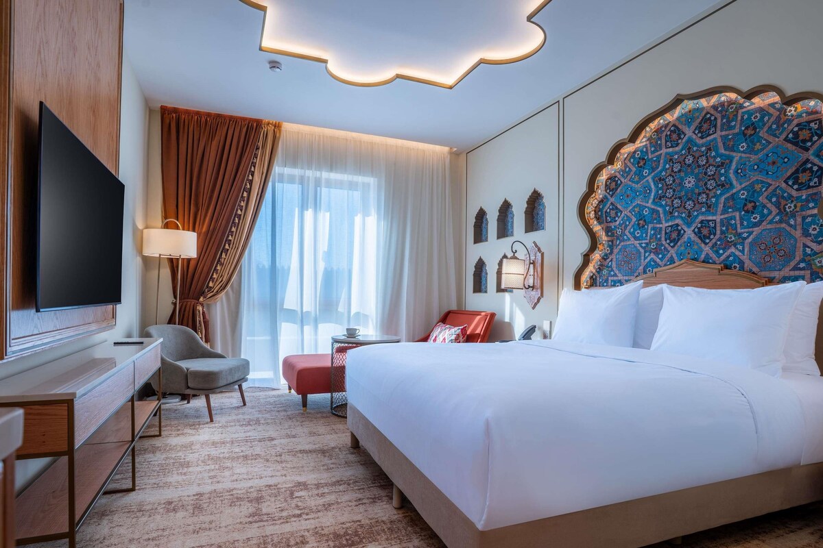 A hotel room with a large bed, a flat-screen TV, a desk with a chair, a small table, and a decorative headboard.