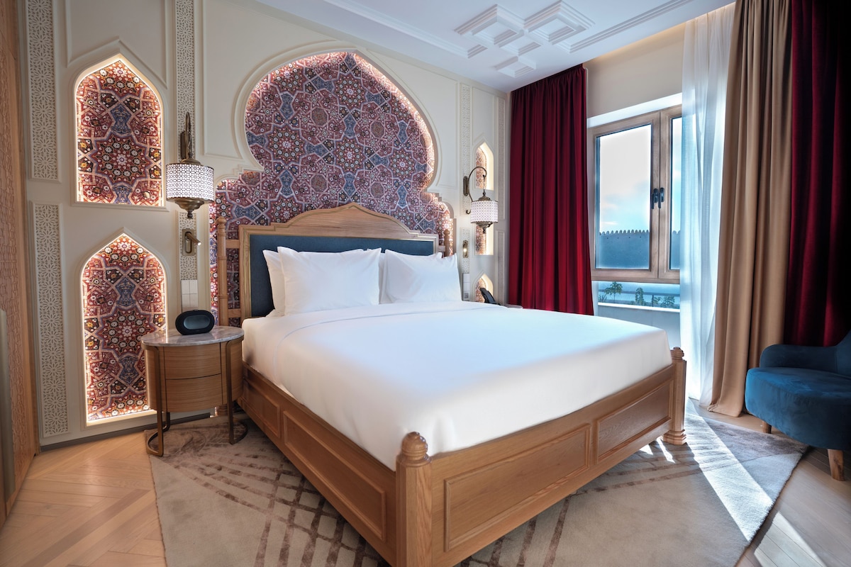 Habitación de hotel con una cama grande, una mesita de noche, una lámpara y vistas al exterior.