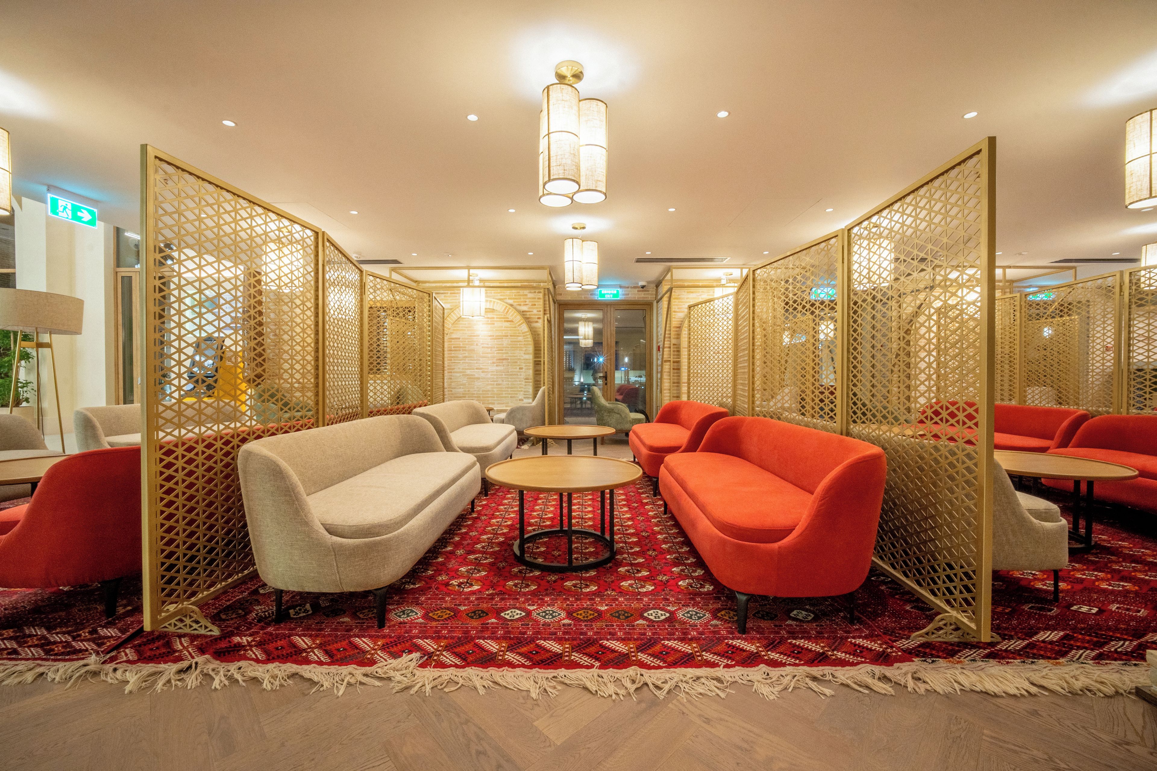 Vestíbulo de hotel con alfombra roja estampada, sofás beige y rojos, una mesita redonda y un separador decorativo.