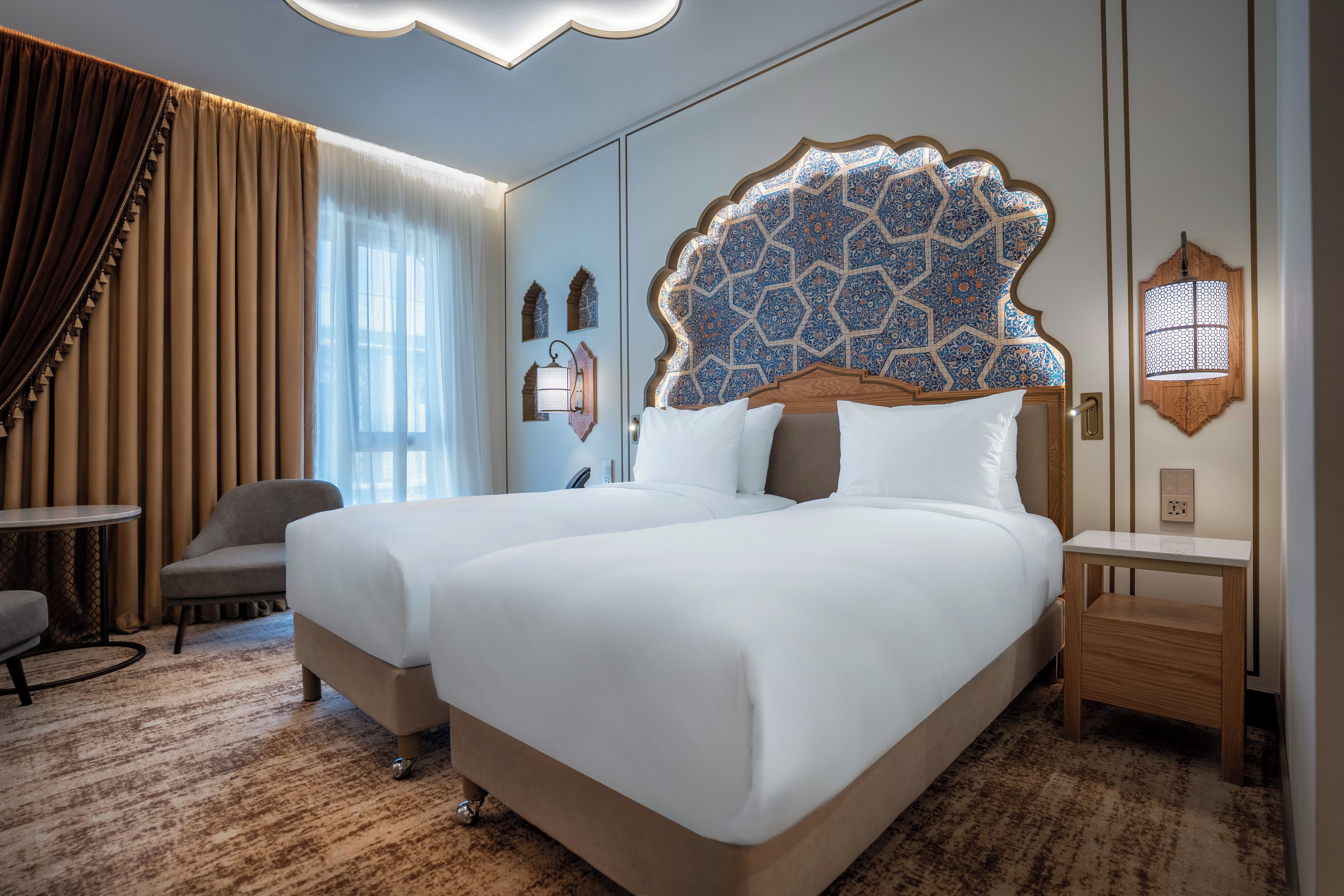 Habitación de hotel con dos camas, una silla, una mesita pequeña y un panel decorativo en la pared.