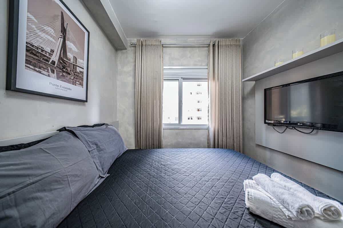 Ein Hotelzimmer mit Bett, Fernseher, Fenster mit Vorhängen und einem gerahmten Bild an der Wand.