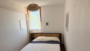 Et soveværelse med en seng, et lille bord, en hængende lampe og et vindue med gardiner.