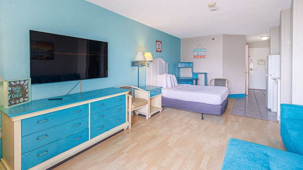 A room with a bed, a desk, a dresser, a television, and a door leading to another room. - Sea Mist 51202 Cozy Ocean View Studio (Myrtle Beach)