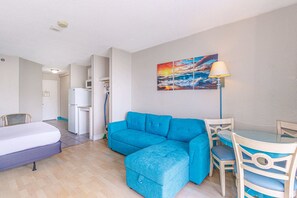 A room with a blue sofa, a bed, a dining table with chairs, a painting on the wall, and a lamp. - The Oceanview | Sea Mist Fifty-One Twenty Oh Two (Myrtle Beach)