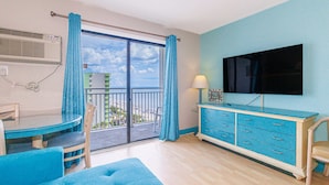 A room with a balcony, a dining table with chairs, a sofa, a TV on a cabinet, and a window with curtains. - Sea Mist 51202 Cozy Ocean View Studio (Myrtle Beach)