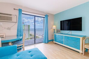 Ein Zimmer mit Balkon, einem Esstisch mit Stühlen, einer Couch, einem Fernseher auf einer Kommode und einem fenster mit Vorhängen.