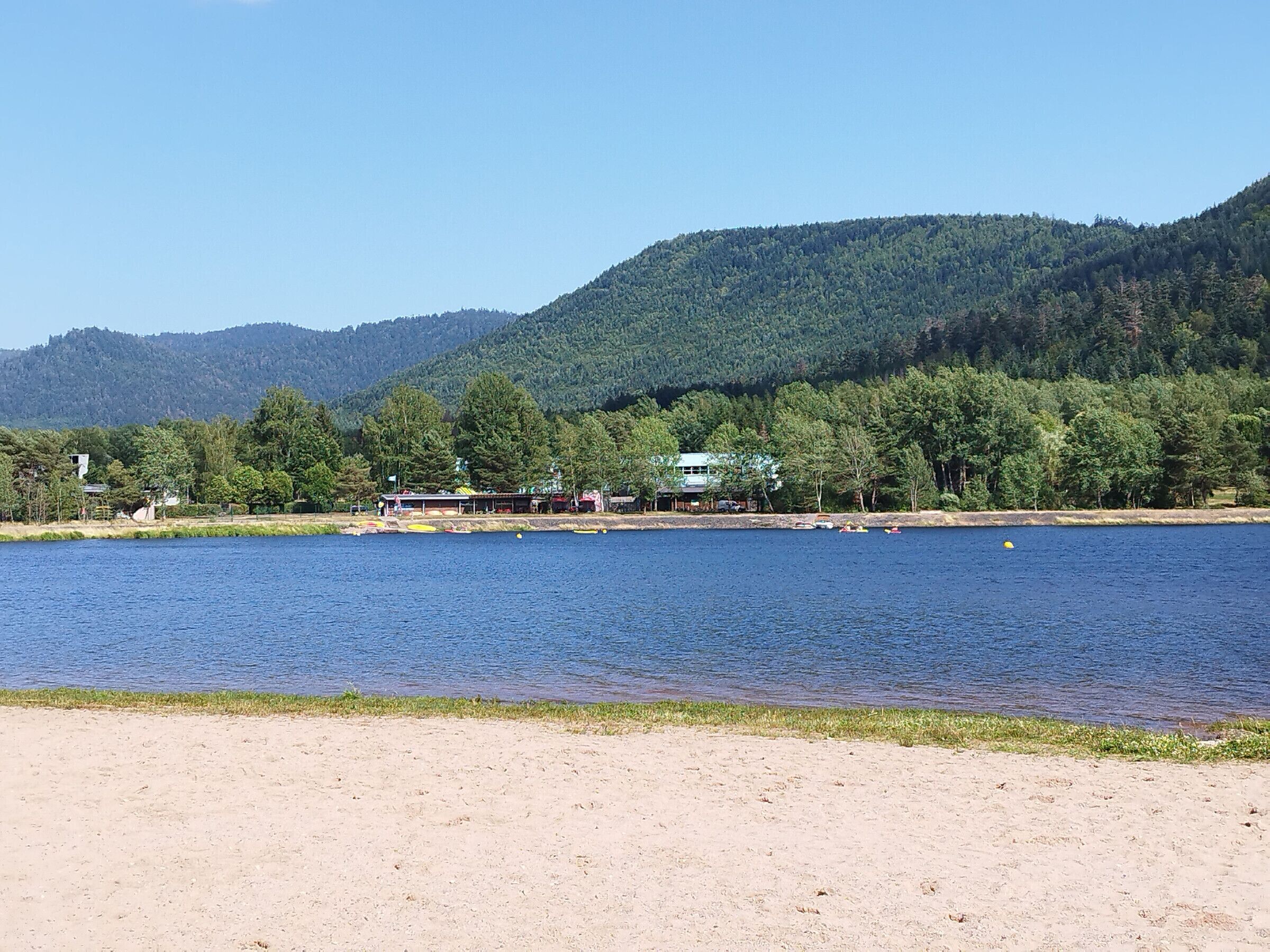 Une vue sur le lac, avec une plage de sable, une rangée de bâtiments et une colline boisée en arrière-plan.